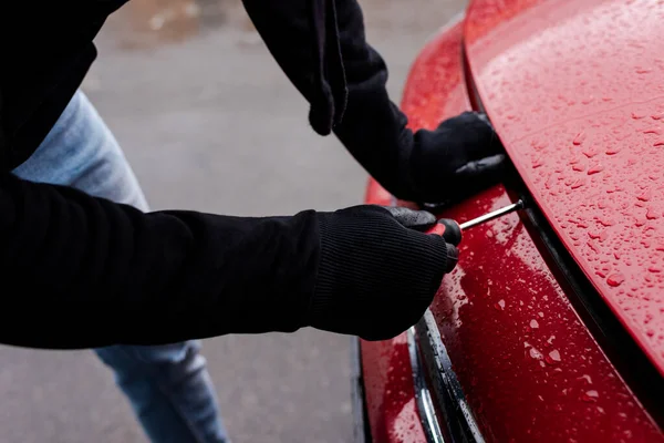 Vista recortada del ladrón en guantes de cuero usando destornillador mientras se abre la capucha del coche en la calle urbana - foto de stock