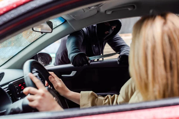 Enfoque selectivo del ladrón en la máscara de la celebración de cuchillo cerca de la mujer sentada en el coche - foto de stock