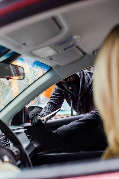 Selective focus of robber in balaclava holding knife and opening car door near driver — Stock Photo