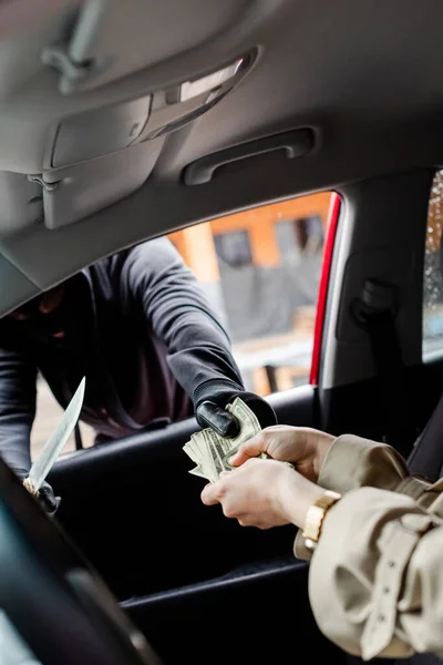 Selective focus of robber in balaclava holding knife and taking dollars from driver in car — Stock Photo