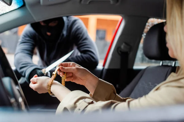 Enfoque selectivo de la mujer quitándose el reloj de pulsera en coche cerca de ladrón con cuchillo - foto de stock