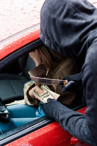Foco seletivo do ladrão segurando faca e dólares perto do motorista assustado no carro — Fotografia de Stock
