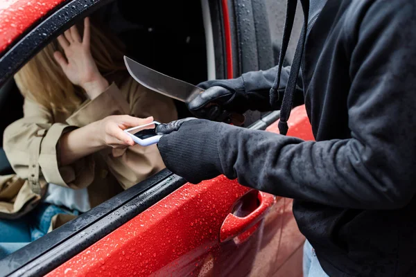 Enfoque selectivo del ladrón sosteniendo el cuchillo mientras toma el teléfono inteligente del conductor asustado en el coche - foto de stock
