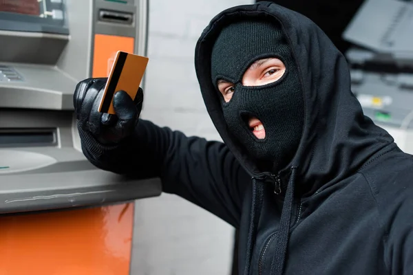 Ladrón sonriente en pasamontañas mirando a la cámara mientras sostiene la tarjeta de crédito cerca del cajero automático - foto de stock