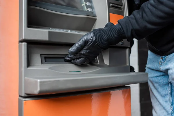 Vista cortada do ladrão em luva de couro segurando a mão perto do dispensador de dinheiro da caixa automatizada — Fotografia de Stock