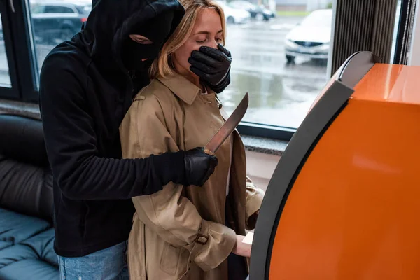 Грабитель в маске держит нож рядом с женщиной с помощью банкомата — стоковое фото