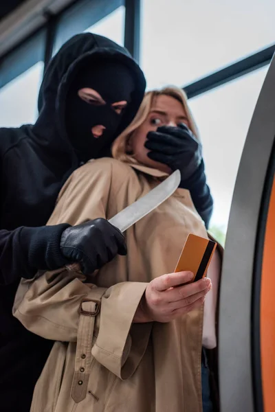 Vista a basso angolo del ladro che tiene il coltello e copre la bocca alla donna spaventata che tiene la carta di credito vicino bancomat — Foto stock