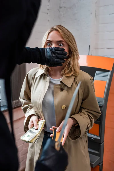 Selective focus of robber with knife covering mouth to scared woman holding credit card and money — Stock Photo