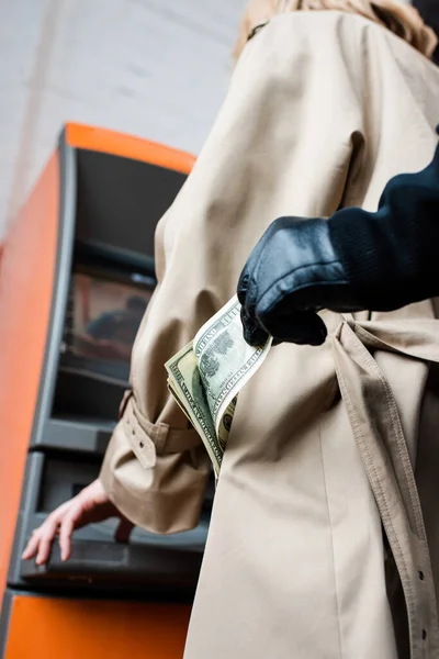 Vista ritagliata di rapinatore in guanto di pelle che prende banconota dollaro dalla tasca del cappotto femminile vicino bancomat — Foto stock