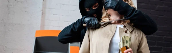 Panoramic crop of robber in balaclava holding knife near female neck during robbery — Stock Photo