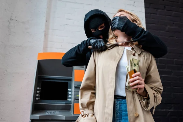 Ladrón en la máscara de la celebración de cuchillo cerca de la mujer con dinero y tarjeta de crédito - foto de stock