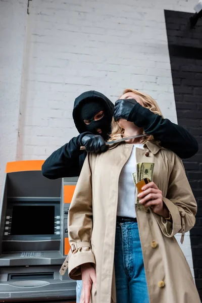 Ladrão segurando faca perto do pescoço da menina segurando dólares e cartão de crédito ao lado ATM — Fotografia de Stock