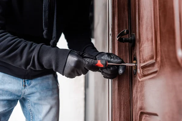 Vista recortada de la rompe-casas en guantes de cuero rompiendo cerradura de la puerta con destornillador - foto de stock