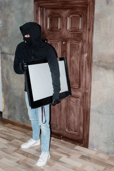 Thief in balaclava and leather gloves holding tv near house door — Stock Photo