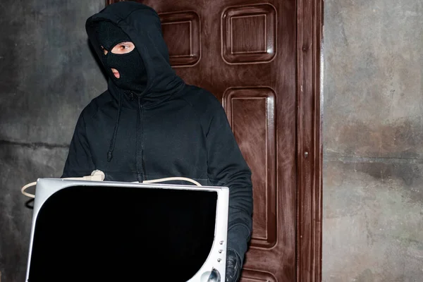 Грабитель в балаклаве держит микроволновую печь во время кражи дома — стоковое фото
