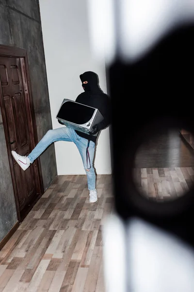 Selective focus of robber in balaclava and leather gloves holding microwave and closing house door — Stock Photo