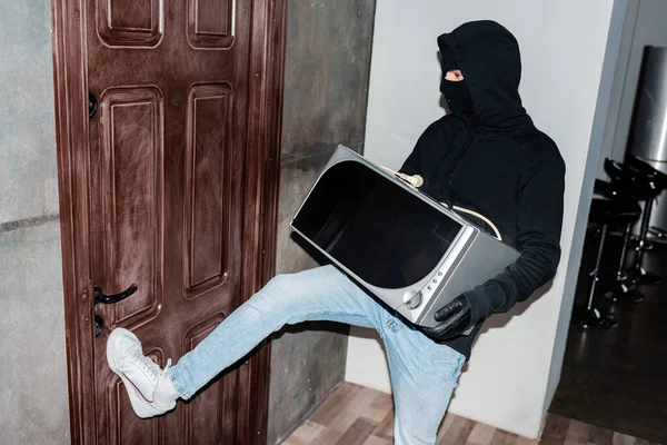 Thief in mask holding microwave and closing house door during stealing — Stock Photo