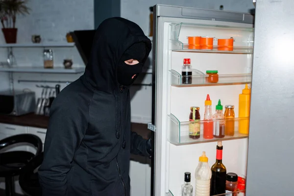 Vista lateral del ladrón en pasamontañas apertura nevera en la cocina - foto de stock