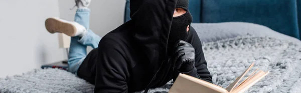 Horizontal crop of robber in balaclava holding book on bed — Stock Photo