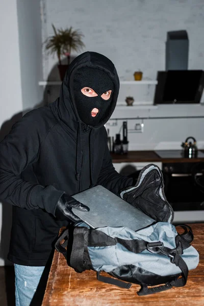 Räuber mit Sturmhaube schaut in Kamera, während er Laptop in Tasche steckt — Stockfoto