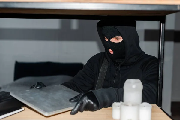 Selective focus of robber in balaclava taking laptop from cupboard — Stock Photo