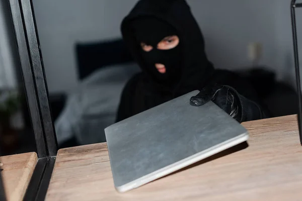 Selective focus of robber in balaclava and leather glove taking laptop from cupboard — Stock Photo