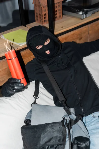 Robber in balaclava holding dynamite near bag with laptop on couch — Stock Photo