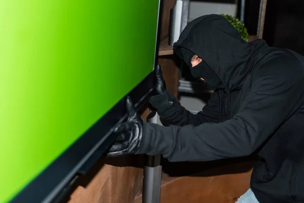 Vue latérale du voleur en masque et gants en cuir tenant la télévision pendant le vol — Photo de stock