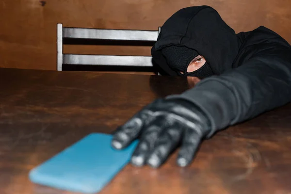 Selective focus of thief in balaclava and leather glove taking smartphone from table — Stock Photo