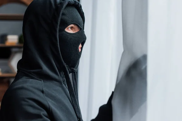 Selective focus of burglar in balaclava standing near curtains — Stock Photo