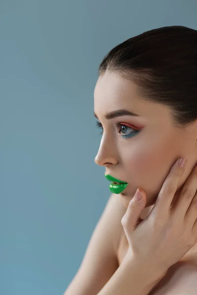 Retrato de mulher bonita nua com lábios verdes, rosa e azul sombra de olhos tocando rosto isolado em azul — Fotografia de Stock