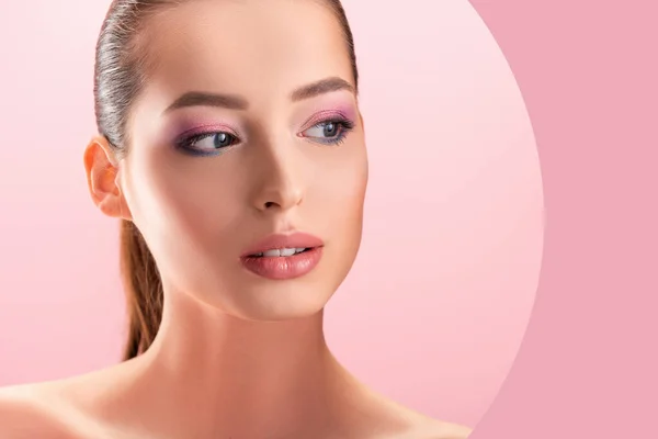 Hermosa mujer desnuda con maquillaje brillante mirando a través de agujero de papel redondo aislado en rosa - foto de stock