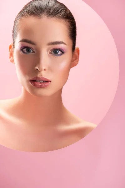 Retrato de bela mulher nua com maquiagem brilhante olhando para a câmera através de buraco de papel redondo isolado em rosa — Fotografia de Stock