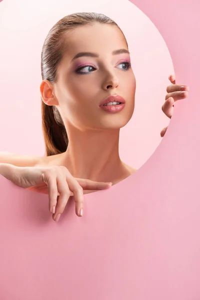 Портрет красивої оголеної жінки з блискучим макіяжем, який дивиться через круглу паперову діру ізольовано на рожевому — стокове фото