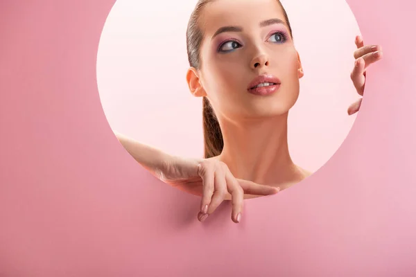 Retrato de bela mulher nua com maquiagem brilhante olhando através de buraco de papel redondo isolado em rosa — Fotografia de Stock