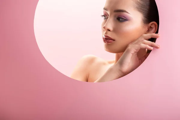 Portrait of beautiful naked woman with shiny makeup looking away through round paper hole isolated on pink — Stock Photo