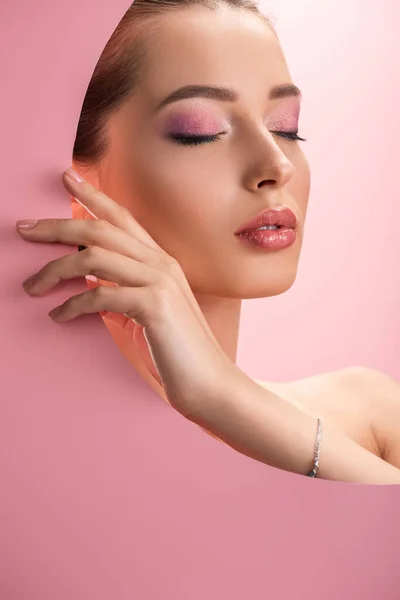 Hermosa mujer desnuda con maquillaje brillante en agujero de papel redondo con los ojos cerrados aislados en rosa - foto de stock
