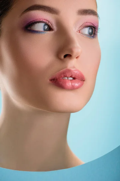 Retrato de mujer hermosa con labios brillantes, sombra de ojos rosa mirando a través de agujero redondo de papel aislado en azul — Stock Photo