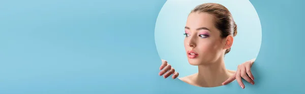 Portrait of beautiful naked woman with glossy lips, pink eyeshadow looking away through paper round hole isolated on blue, panoramic shot — Stock Photo