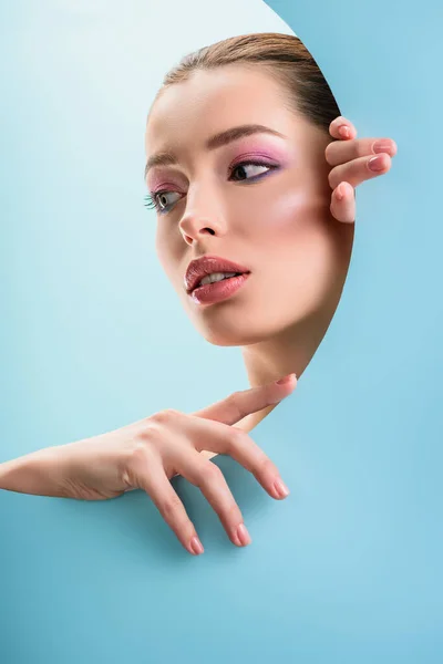 Retrato de mujer hermosa con labios brillantes, sombra de ojos rosa mirando a través de agujero redondo de papel aislado en azul - foto de stock