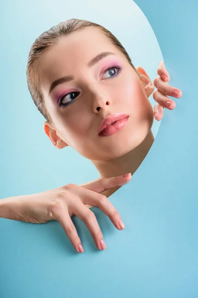 Retrato de mujer desnuda hermosa con labios brillantes, sombra de ojos rosa mirando a través de agujero redondo de papel aislado en azul — Stock Photo