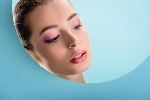 Porträt einer schönen jungen Frau mit glänzenden Lippen, rosa Lidschatten, der durch ein rundes, auf blau isoliertes Loch aus Papier nach unten blickt — Stockfoto