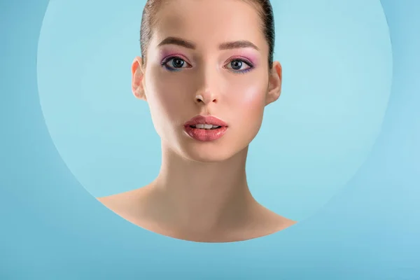 Retrato de mulher nua bonita com lábios brilhantes, sombra de olhos rosa olhando através de papel buraco redondo isolado em azul — Fotografia de Stock