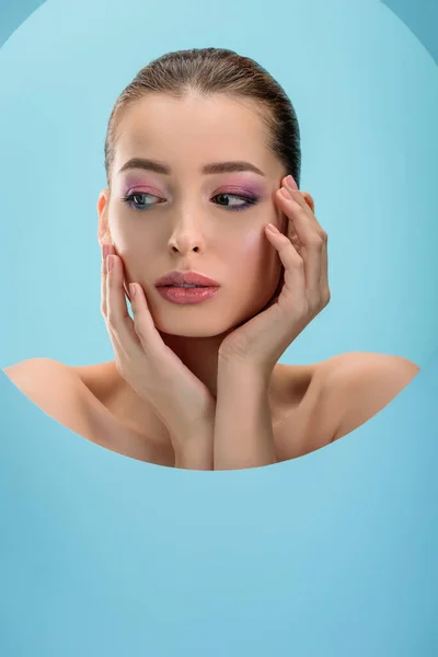 Retrato de mulher nua bonita com lábios brilhantes, sombra de olhos rosa e mãos no rosto em papel buraco redondo isolado em azul — Fotografia de Stock