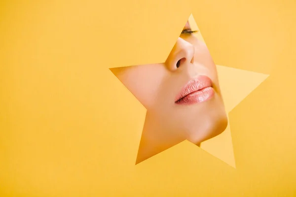 Vista recortada de la hermosa mujer con labios brillantes en el agujero en forma de estrella de papel aislado en amarillo - foto de stock