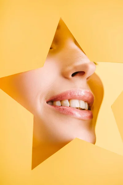 Vista recortada de hermosa mujer con labios brillantes en el agujero en forma de estrella de papel sonriendo aislado en amarillo - foto de stock