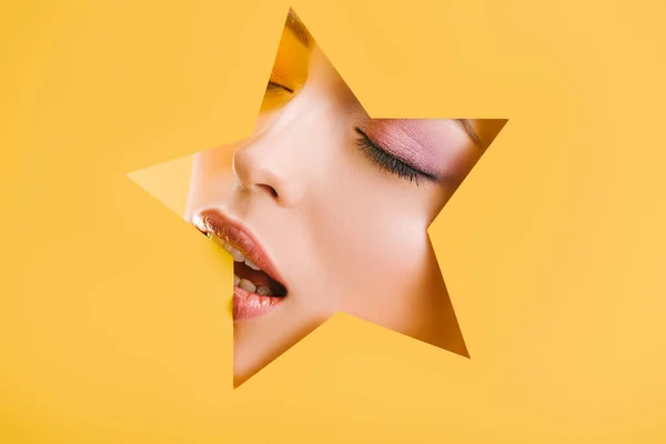 Retrato de mujer hermosa con maquillaje brillante en el agujero en forma de estrella de papel con los ojos cerrados y la boca abierta aislada en amarillo - foto de stock