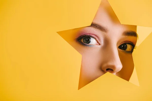 Portrait of beautiful woman with shiny makeup in paper star shaped hole looking at camera isolated on yellow — Stock Photo