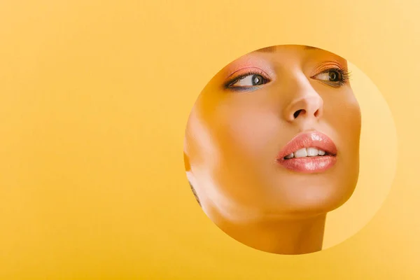 Portrait of beautiful woman with shiny makeup in paper round hole looking away isolated on yellow — Stock Photo