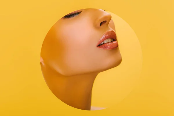 Partial view of beautiful woman with shiny makeup in paper round hole with closed eyes isolated on yellow — Stock Photo